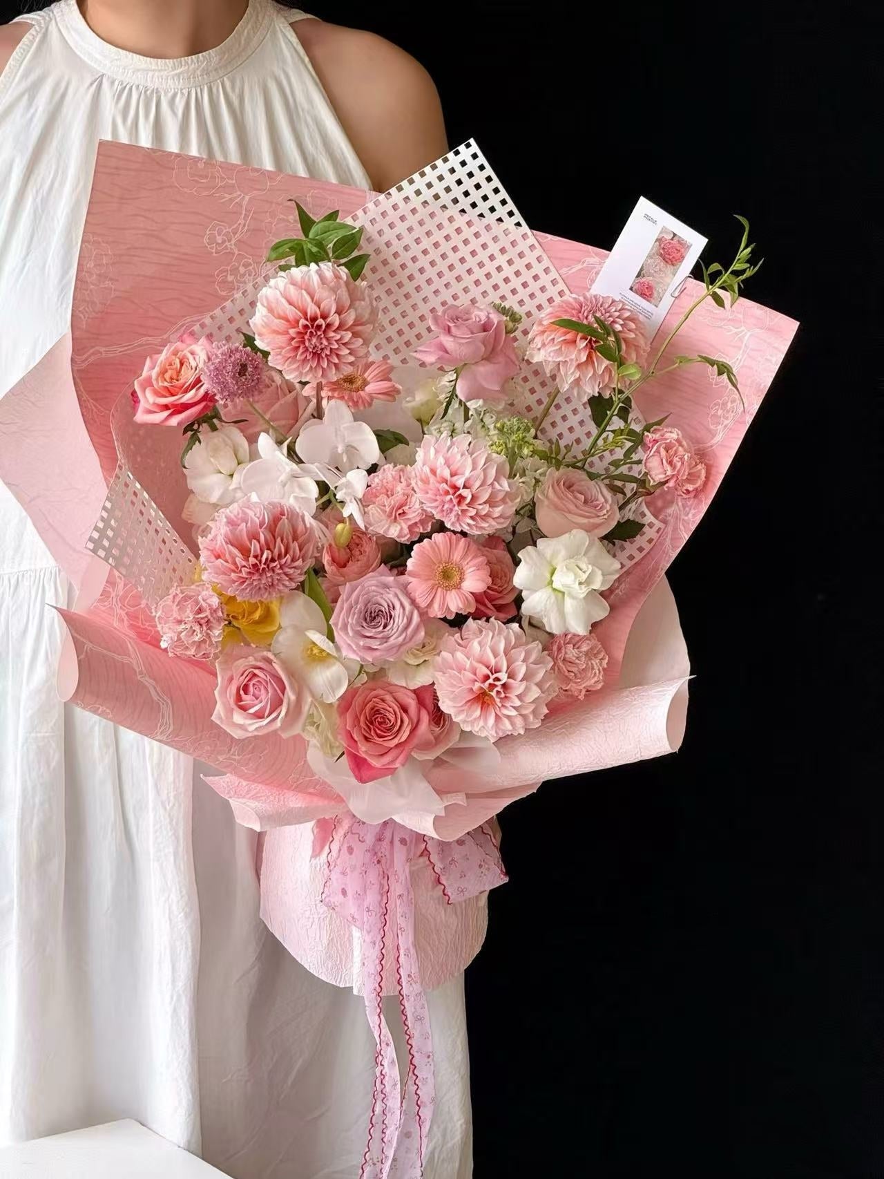 A bouquet of pink and white flowers wrapped in pink paper, held by a person wearing a white sleeveless dress against a black background.